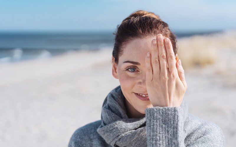 Eine junge lächelnde Frau am Strand hält sich ein Auge zu.