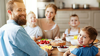 Eine junge Familie frühstückt zusammen am Esstisch. Mutter, Vater und drei Kinder lächeln in die Kamera.